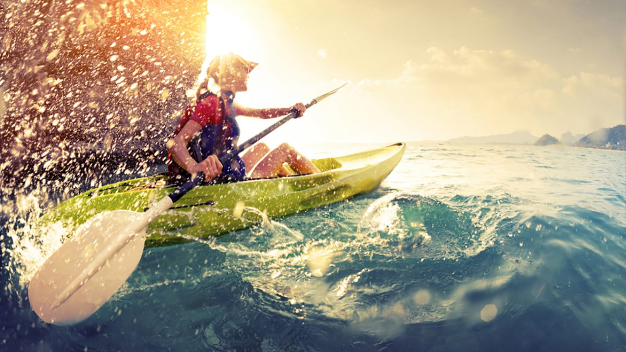 Young lady paddling hard the kayak with lots of splashes near the cliff at sunny day